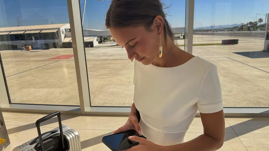 Žena stojí u vysokého prosvětleného okna v příletové hale letiště, dívá se do telefonu a vedle ní stojí malý kufr
