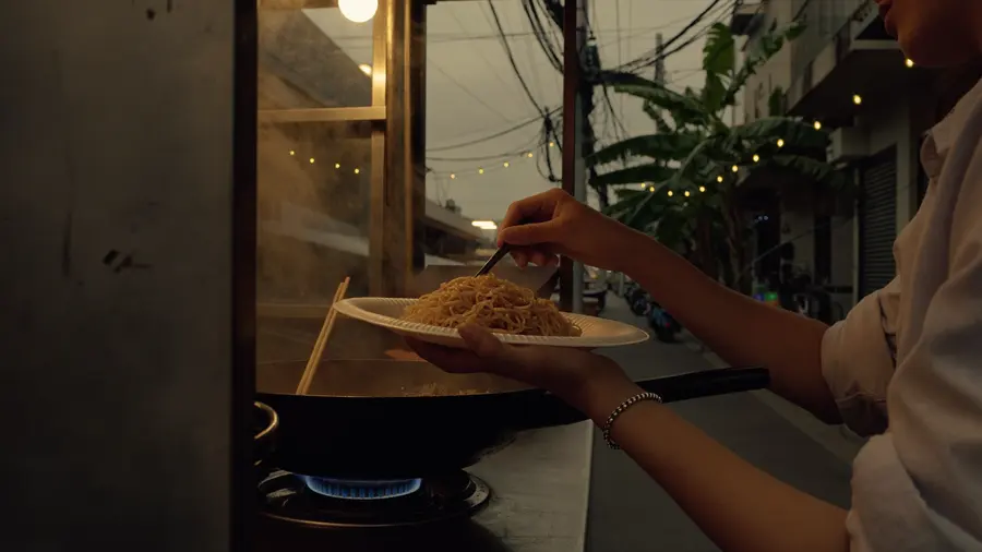 Una persona in viaggio ritira la sera un piatto di noodle dal bancone di un piccolo carretto di cibo di strada