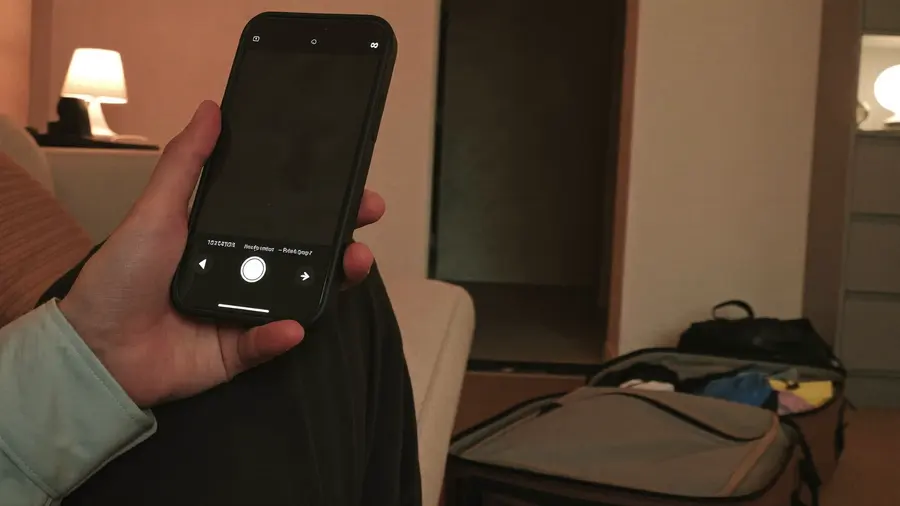 Una persona seduta sul divano nella luce calda di una lampada la sera, con il telefono in mano e un bagaglio a mano aperto per terra accanto a lei