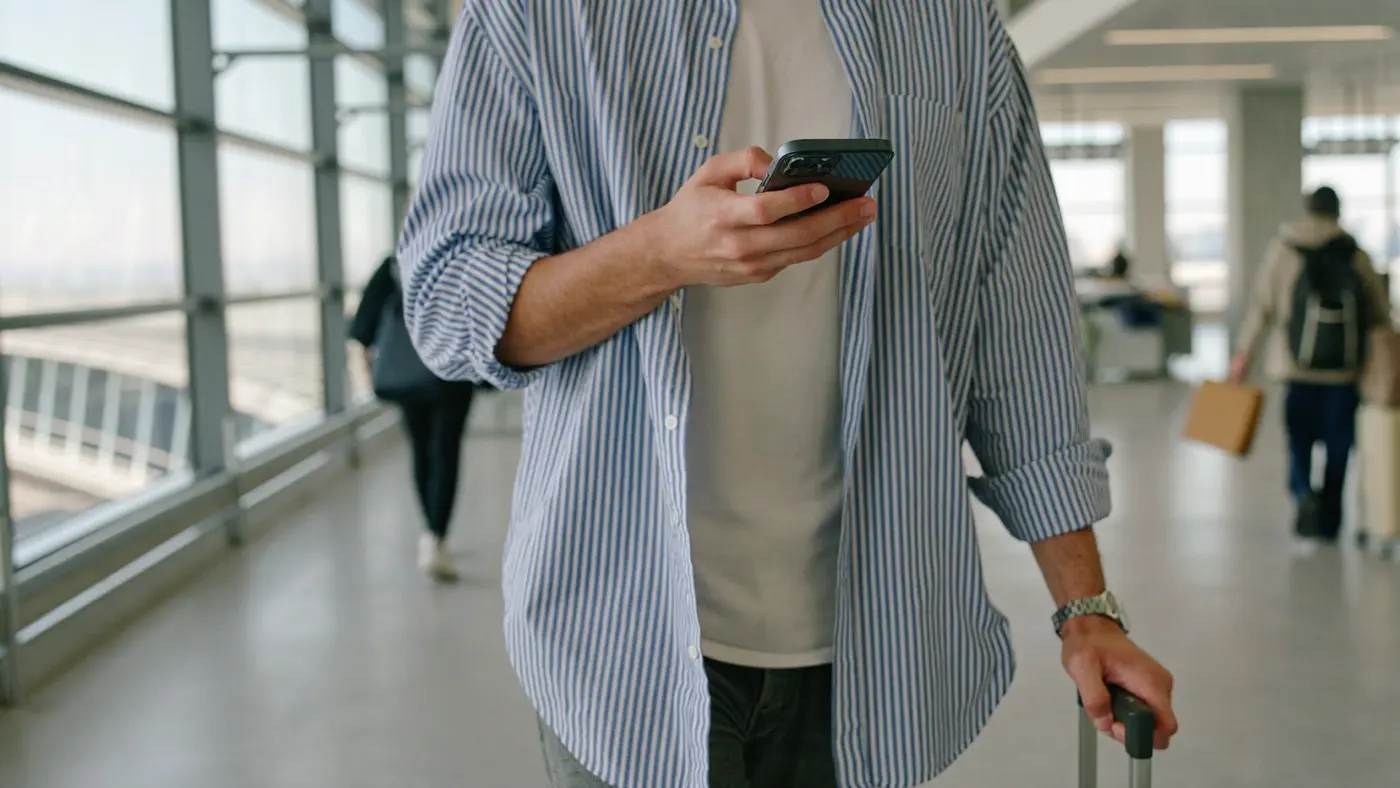 Una persona attraversa con una valigia una luminosa sala arrivi guardando un telefono tenuto all'altezza del petto