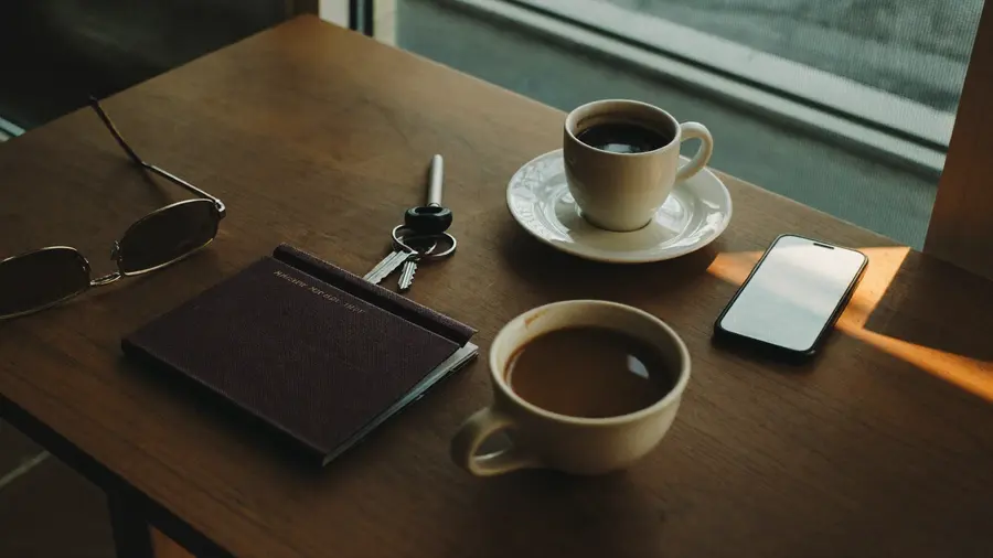 Un tavolo di legno vicino a una finestra con un passaporto, occhiali da sole, chiavi e un telefono appoggiato a faccia in giù accanto a una tazza di caffè