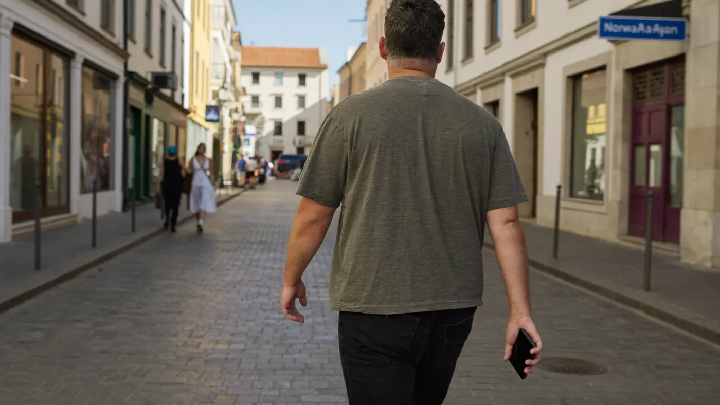 Cestující kráčí sluncem zalitou uličkou starého města, telefon drží volně v jedné ruce a dívá se vzhůru na budovy