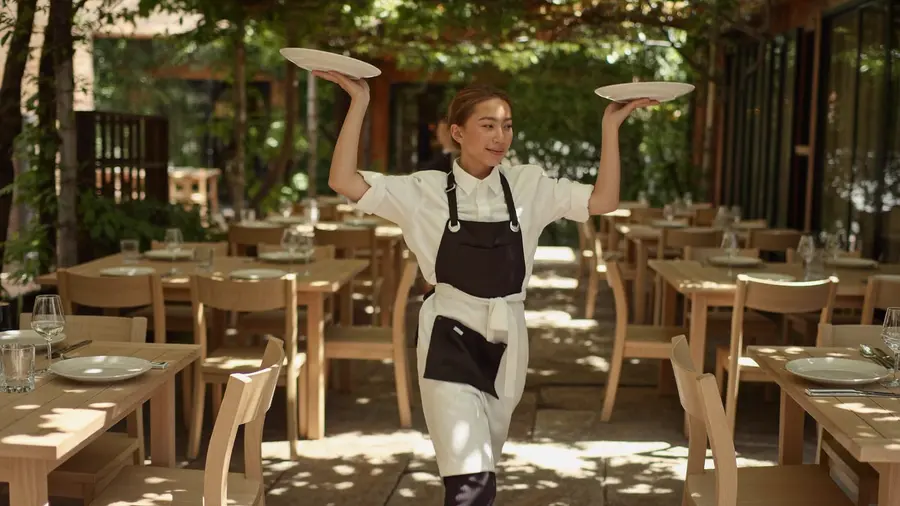 Servírka nese dva talíře přes sluncem zalitou venkovní terasu restaurace
