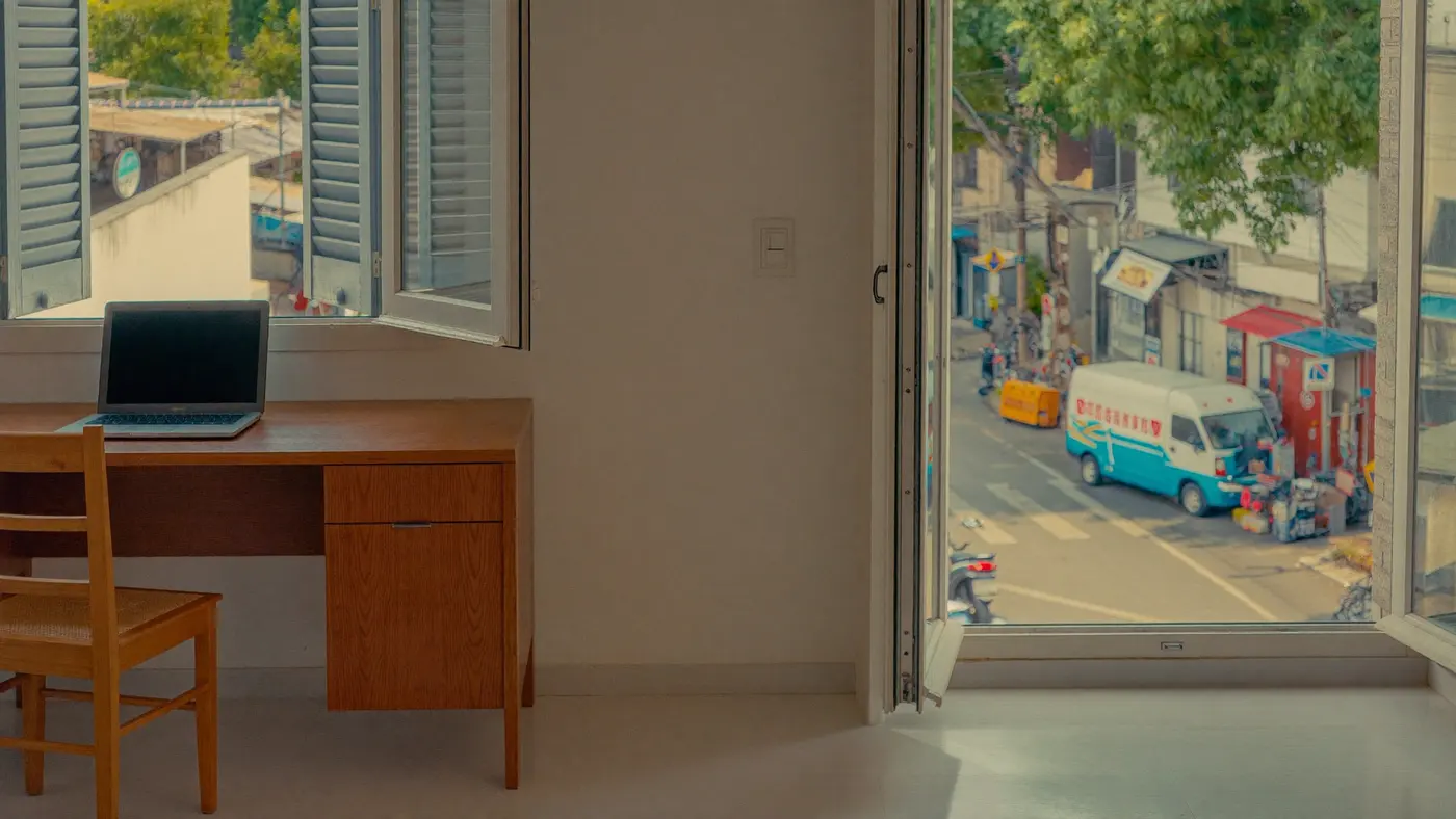Eine helle Mietwohnung mit einem Schreibtisch neben hohen offenen Fenstern mit Blick hinunter auf eine belebte sommerliche Straße