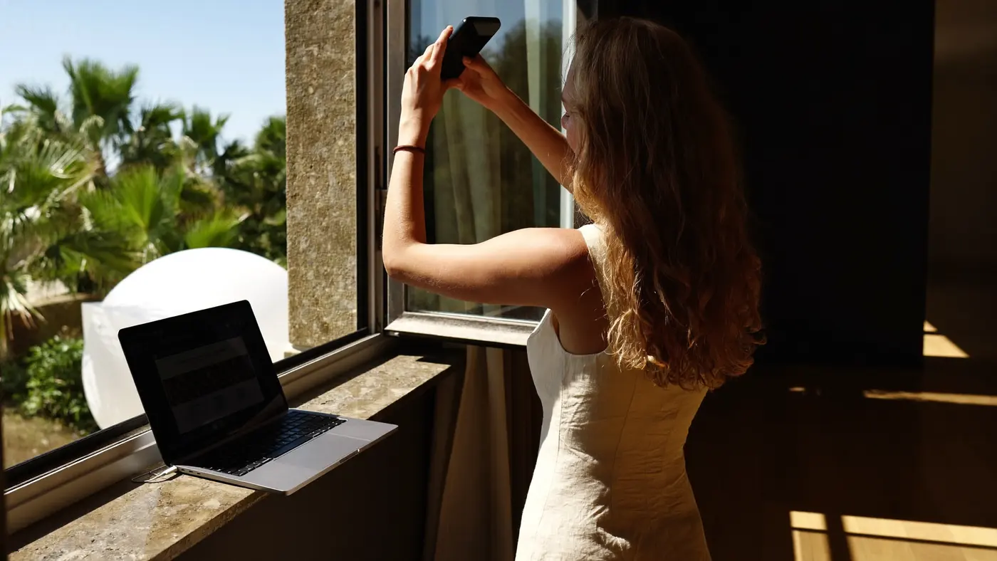 Žena v prázdném prosluněném pokoji zvedá telefon k otevřenému oknu, vedle na parapetu opřený notebook