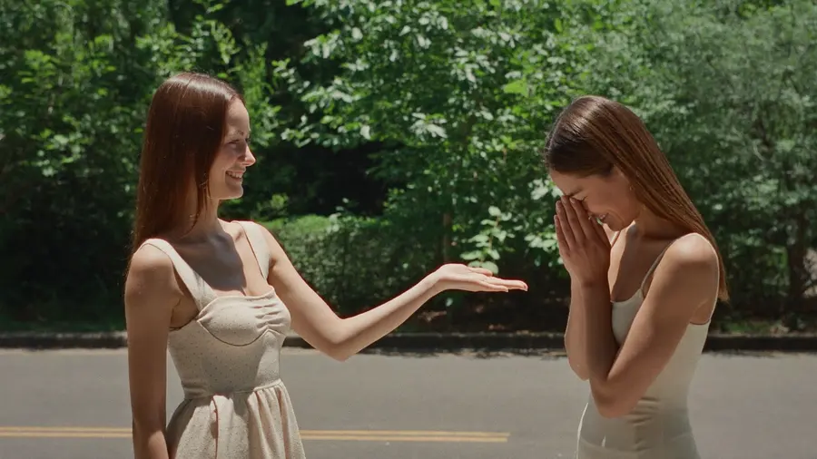 Deux femmes se saluent sur un trottoir ensoleillé, l'une tend la main tandis que l'autre a déjà amorcé une légère inclinaison