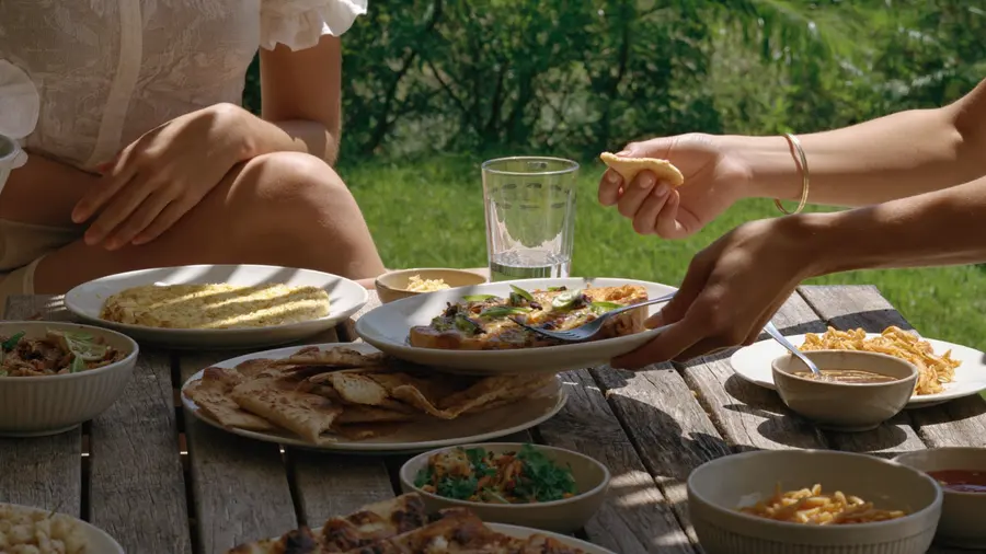 Des mains se croisent au-dessus d'une table de plats partagés sur une terrasse ombragée