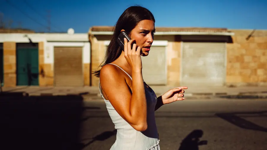 Mladá žena rychle kráčí prázdnou prosluněnou ulicí v ostrém letním světle, telefon u ucha