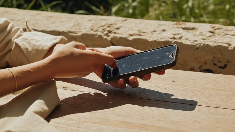 Ženské ruce drží poškrábaný starší telefon se zhasnutým displejem nad sluncem prosvíceným dřevěným stolem